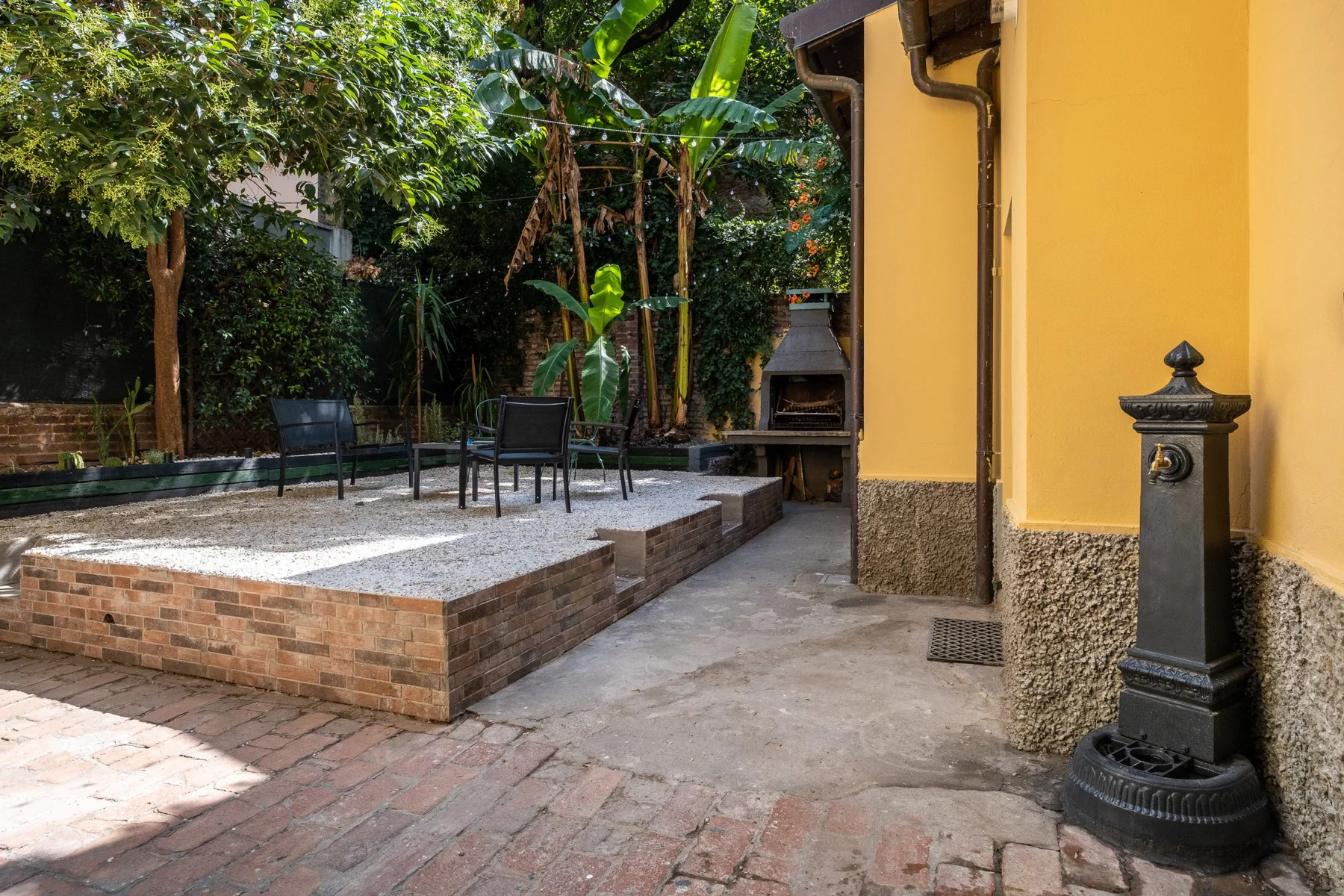 Un patio ajardinado con asientos al aire libre, plantas frondosas y un edificio amarillo.