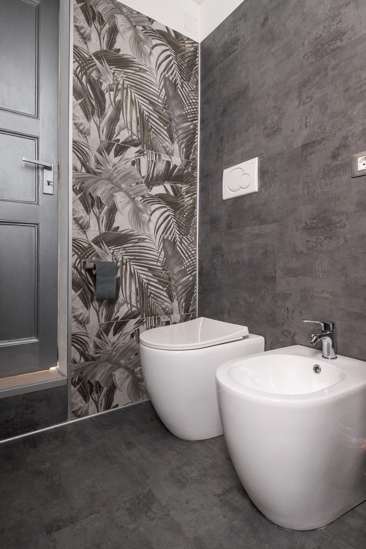 Un baño con inodoro y bidé junto a una pared con papel pintado de hojas tropicales.