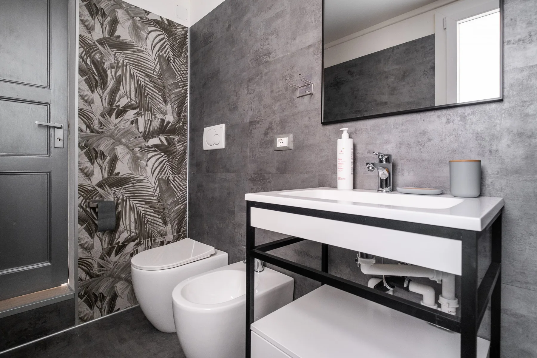 Un baño moderno con lavabo blanco, inodoro y una pared con estampado de hojas.