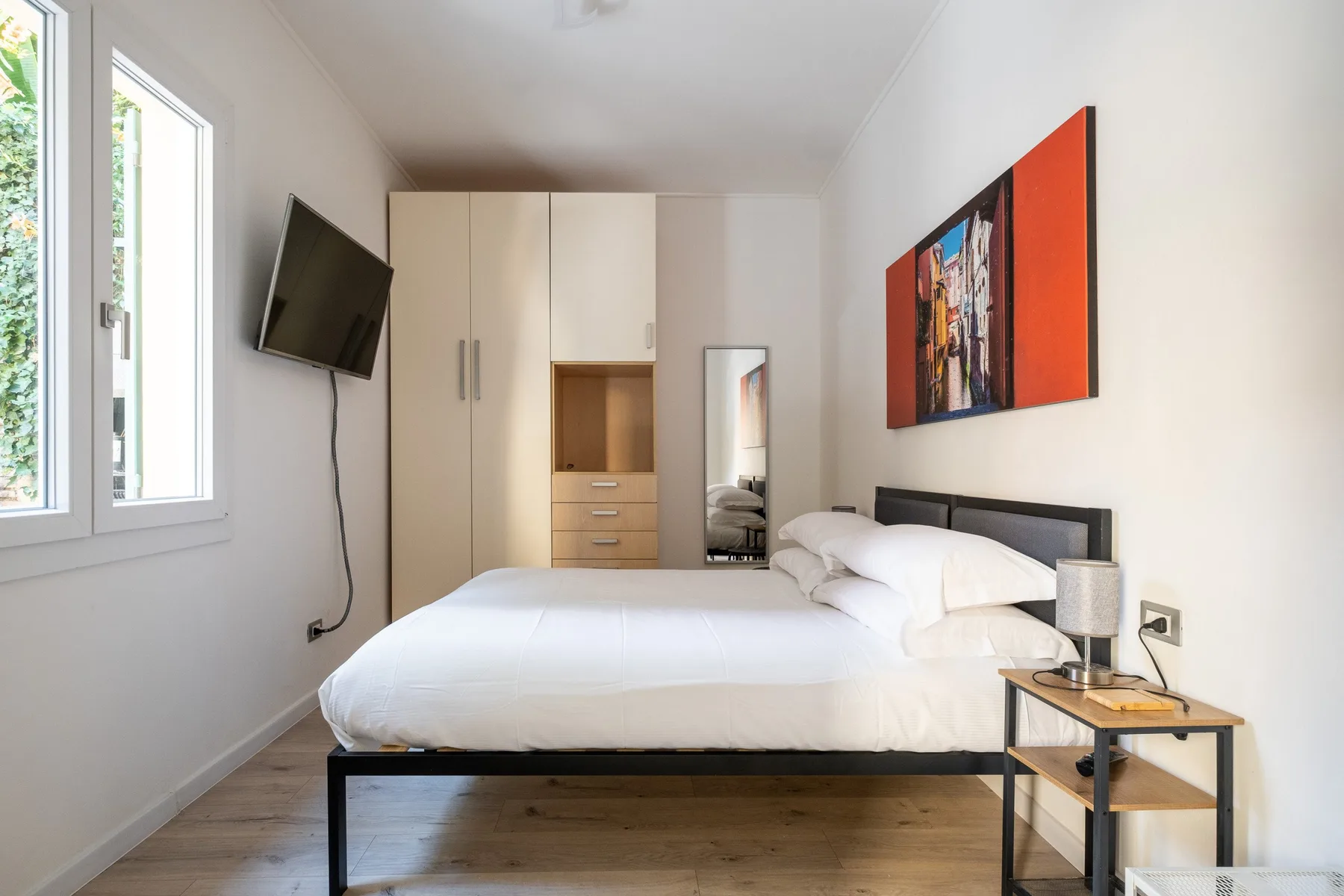 A bright bedroom with a large bed, wardrobe, and wall-mounted television.