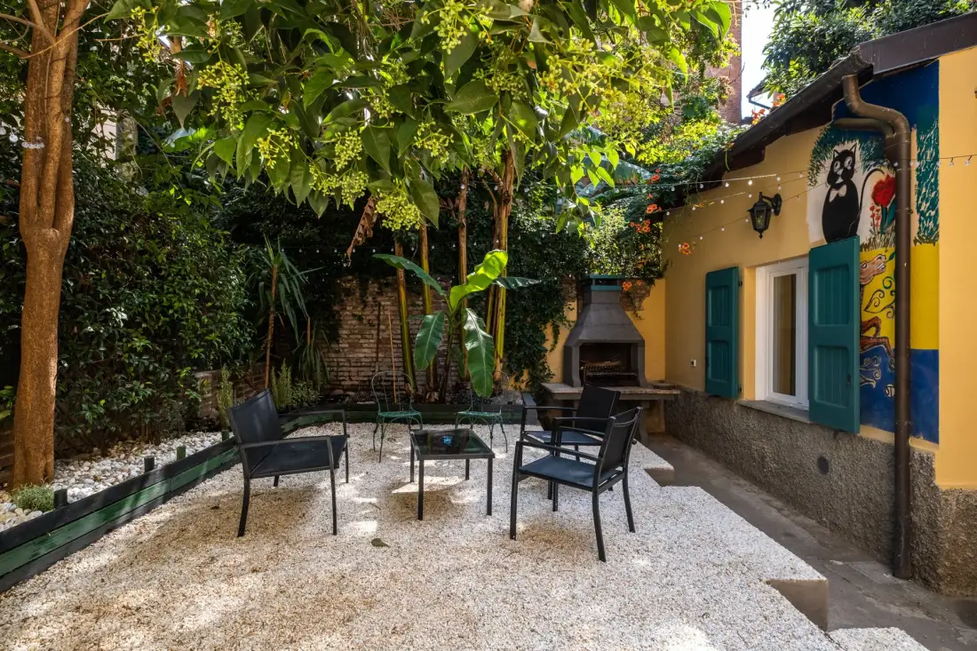 Un patio exterior frondoso con asientos y una barbacoa junto a un edificio colorido.