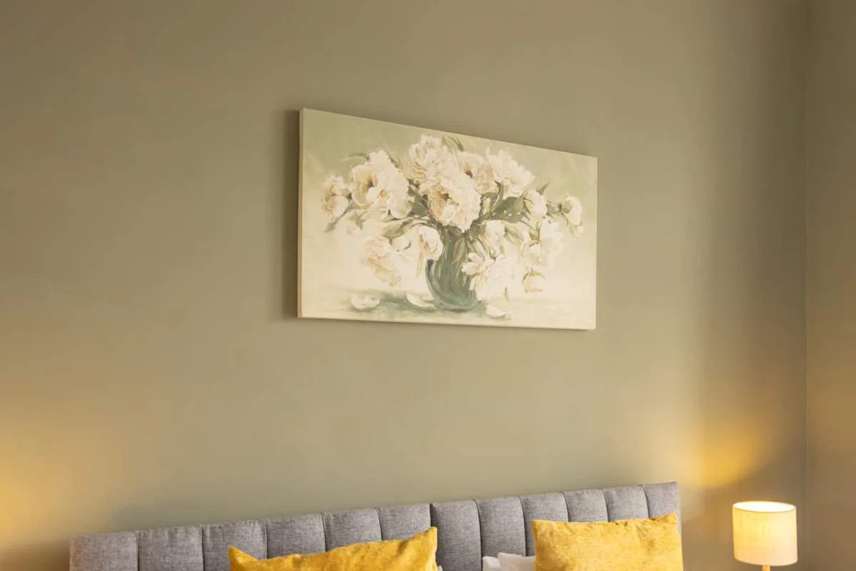 A bedroom seating area has a gray upholstered headboard and floral wall art.