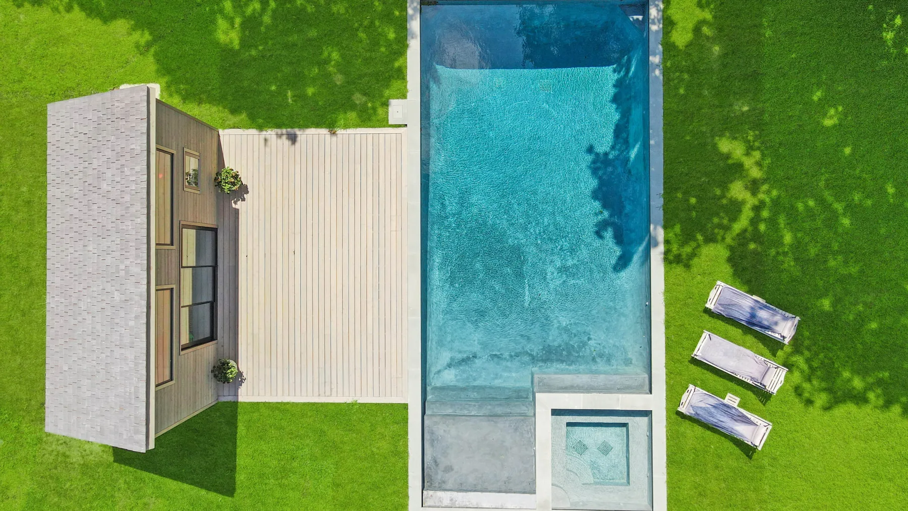 An aerial view shows a swimming pool beside a house and three loungers on the lawn.