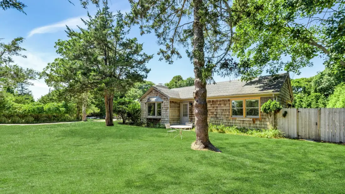 A small house sits beyond a broad green lawn beneath leafy trees.