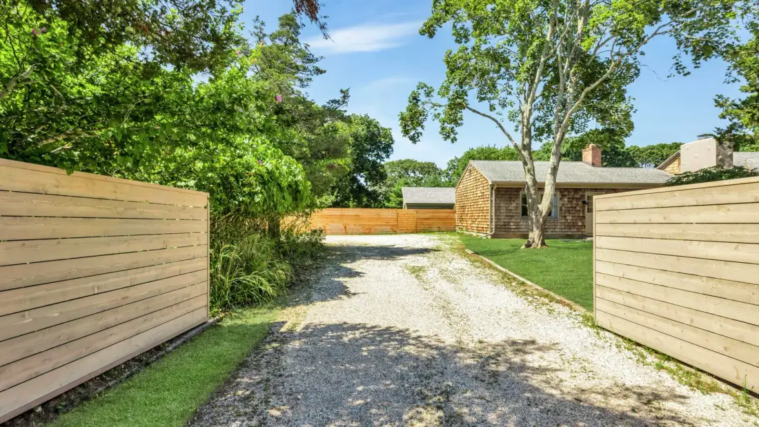 A gravel driveway runs between wooden fences toward a small outbuilding.