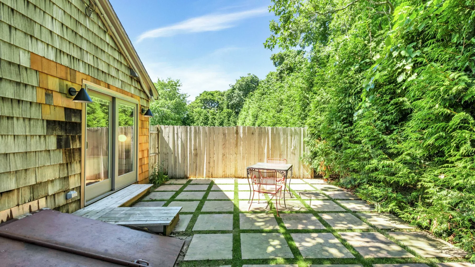 A paved outdoor patio beside a wooden-sided building, with trees and shrubs along the edge.