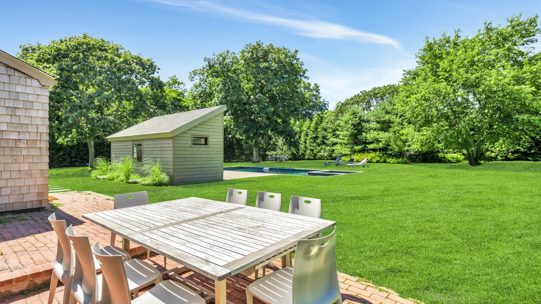 A backyard patio with an outdoor dining table beside a broad lawn and trees.