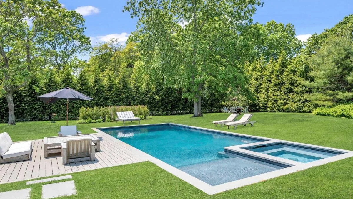 An outdoor swimming pool sits beside a paved seating area in a leafy garden.