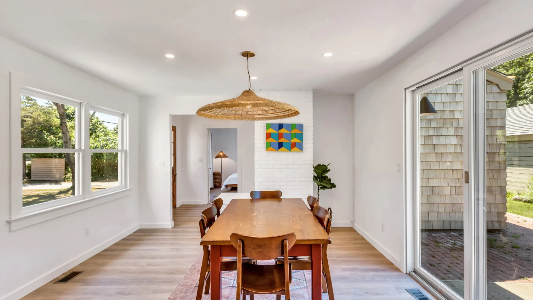 A bright dining room features a wooden table beneath a woven pendant light.