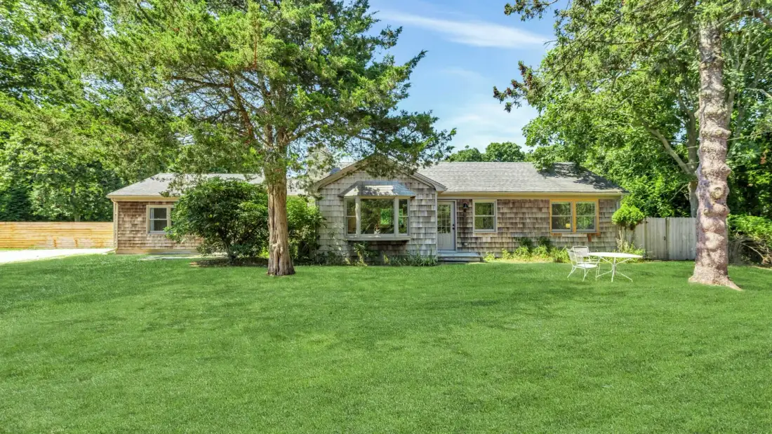 A single-story house stands behind a broad green lawn and mature trees.
