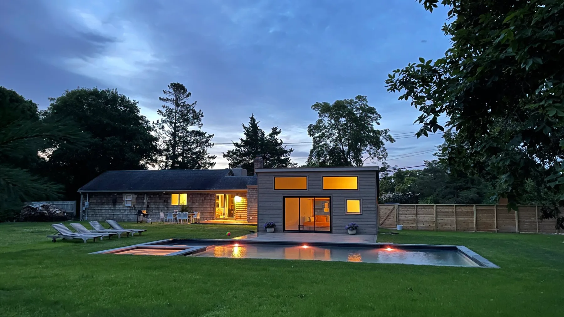 A modern house and illuminated swimming pool sit in a grassy yard at dusk.