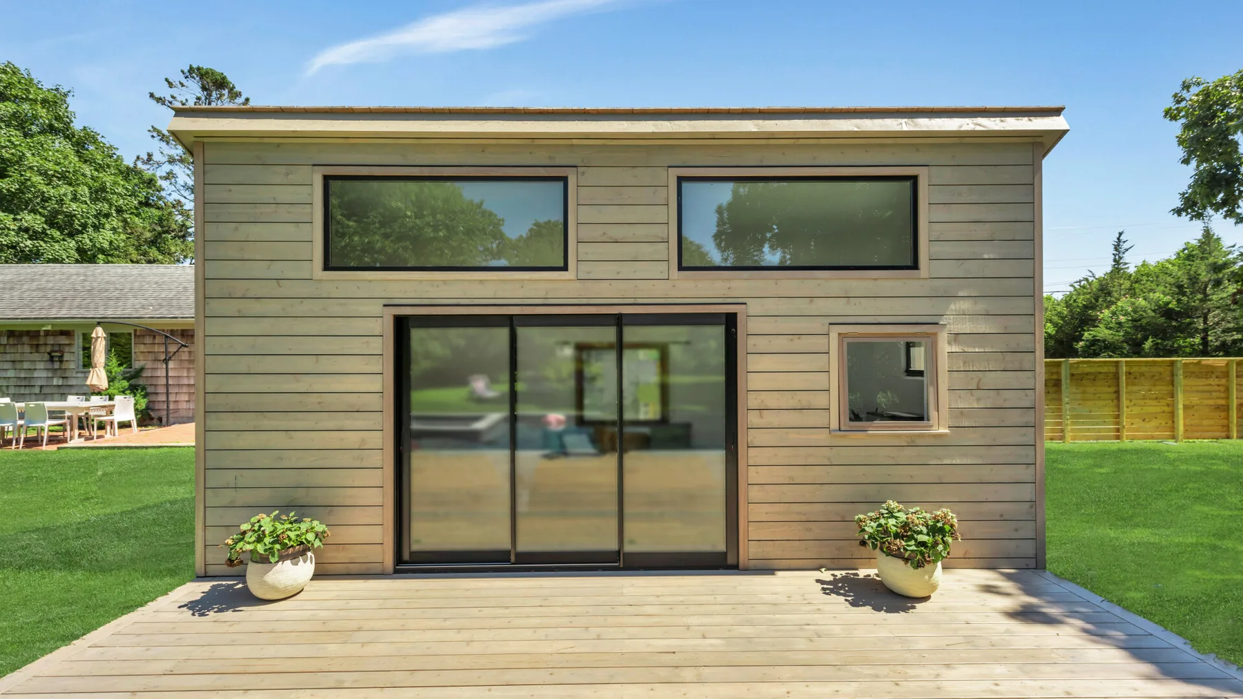 A modern small house with wide windows opens onto a wooden deck and green lawn.