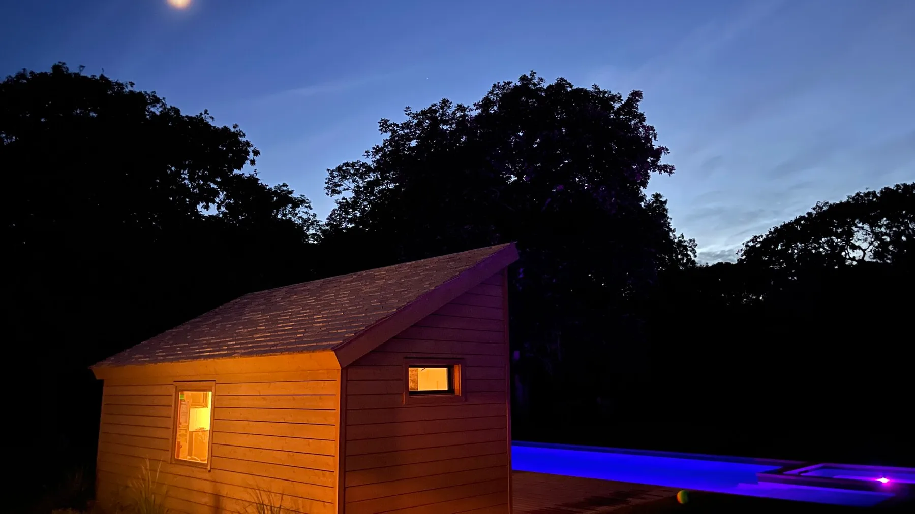A small wooden cabin glows beside a blue-lit pool at dusk.