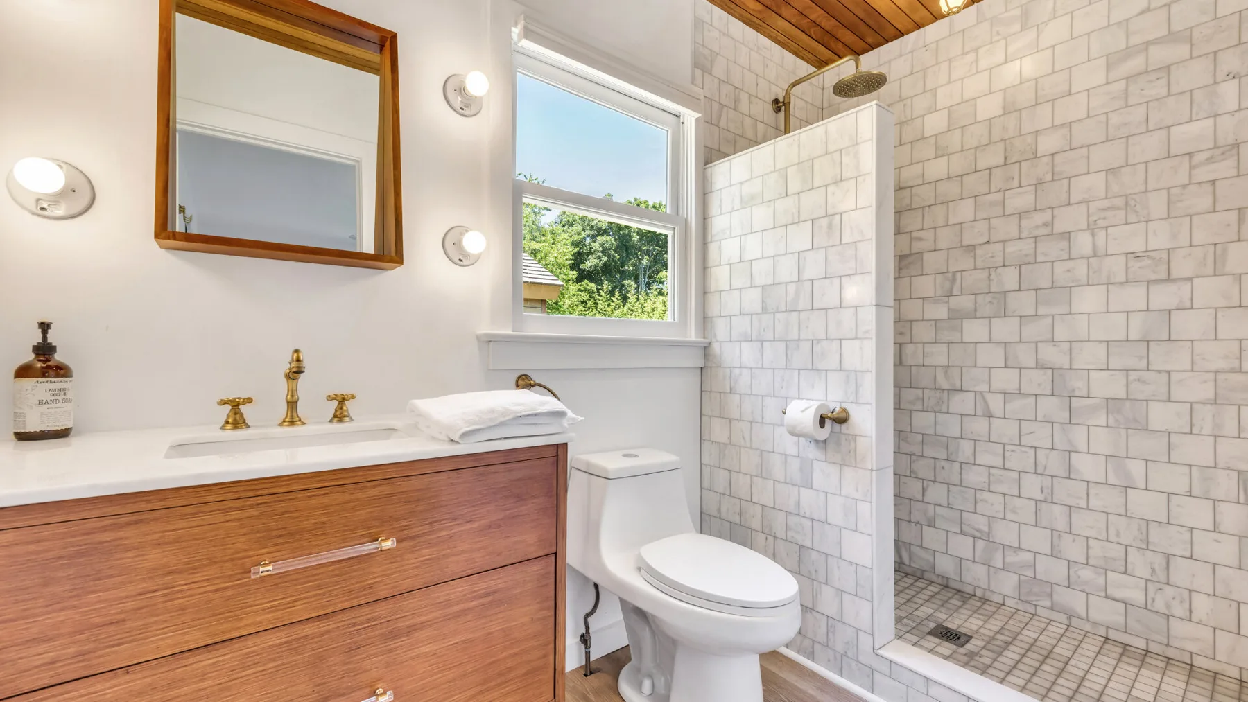 A bright bathroom with a wood vanity, toilet, and tiled shower.