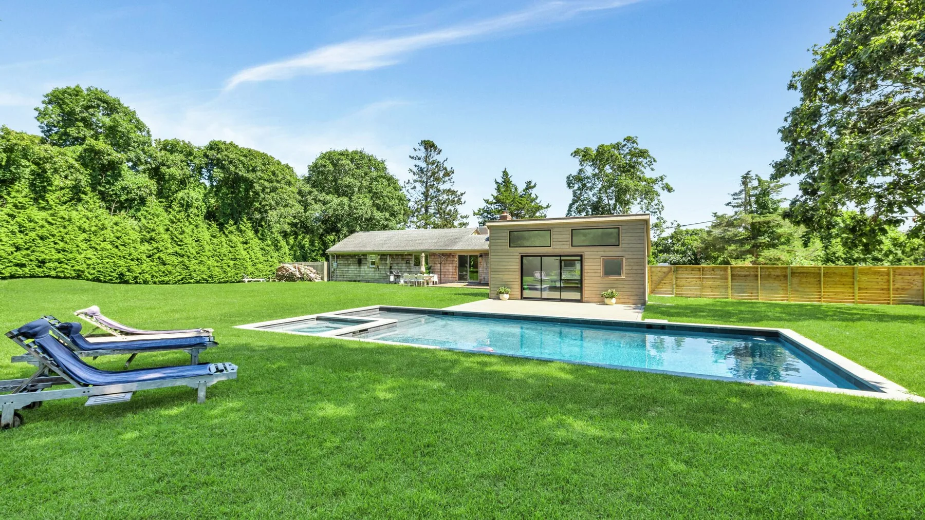A swimming pool and lounge chairs sit in a grassy yard beside a modern house.