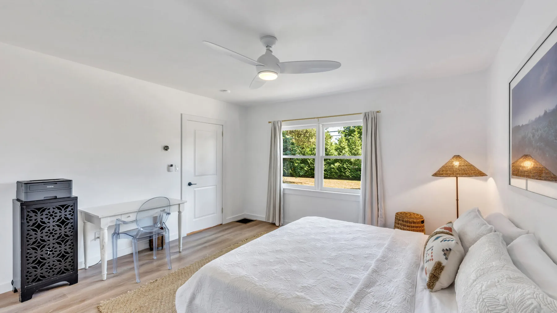 A bright bedroom with a bed, desk, and window overlooking greenery.
