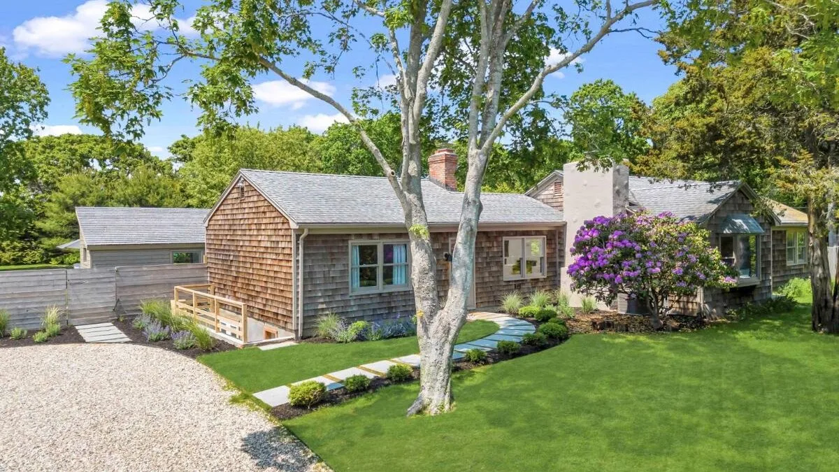 A cottage-style house sits beside a green lawn, with trees and flowering shrubs.