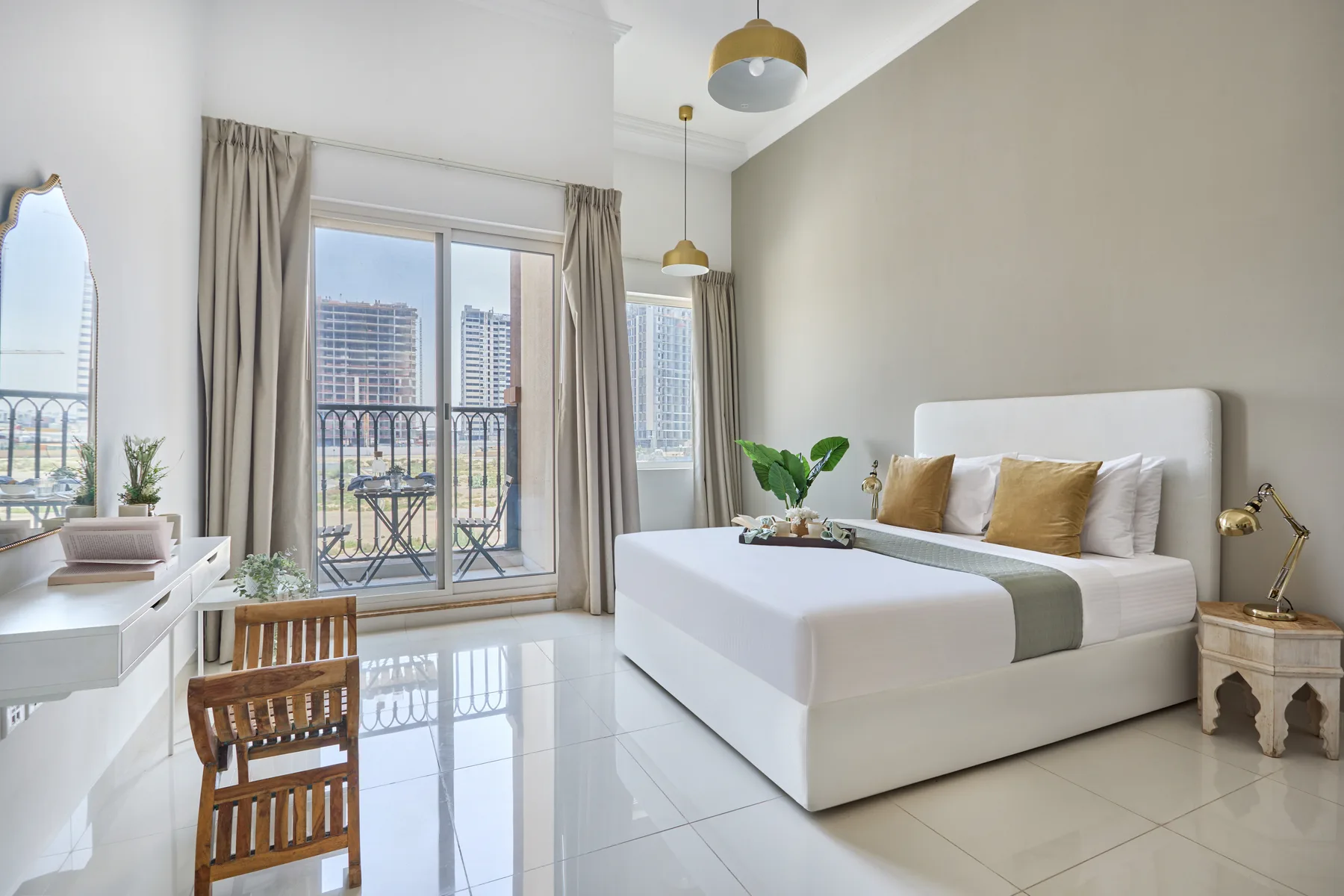 Un dormitorio luminoso con una cama blanca y grandes ventanales frente a edificios cercanos.