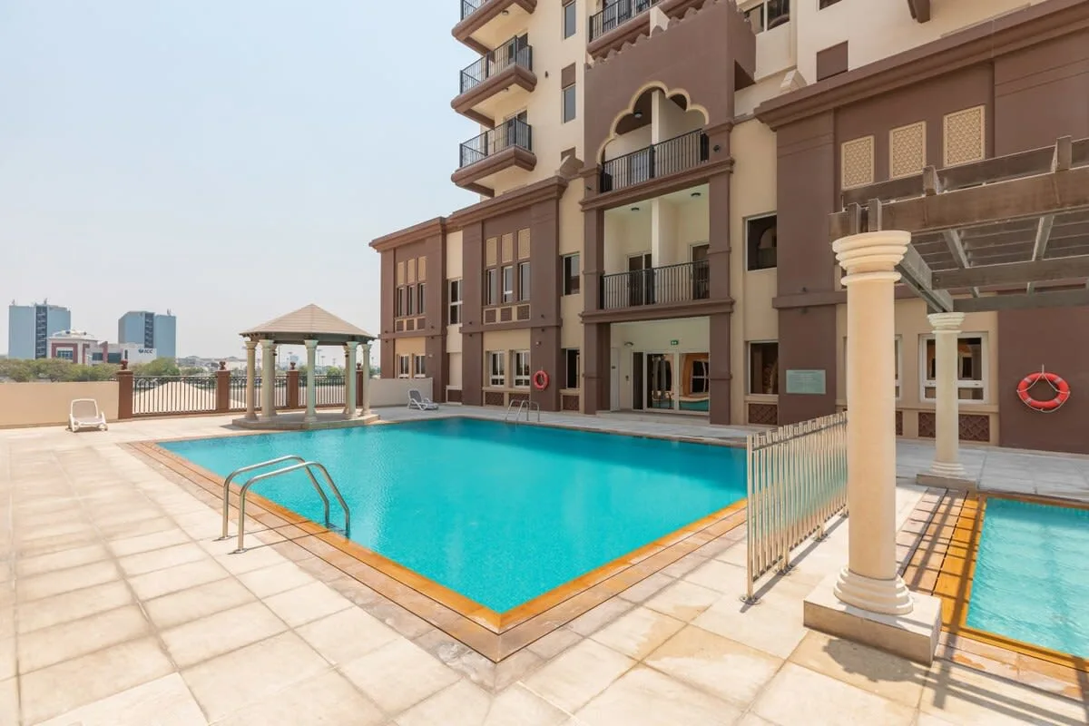 Una piscina al aire libre está junto a un edificio de varios pisos y una terraza pavimentada.
