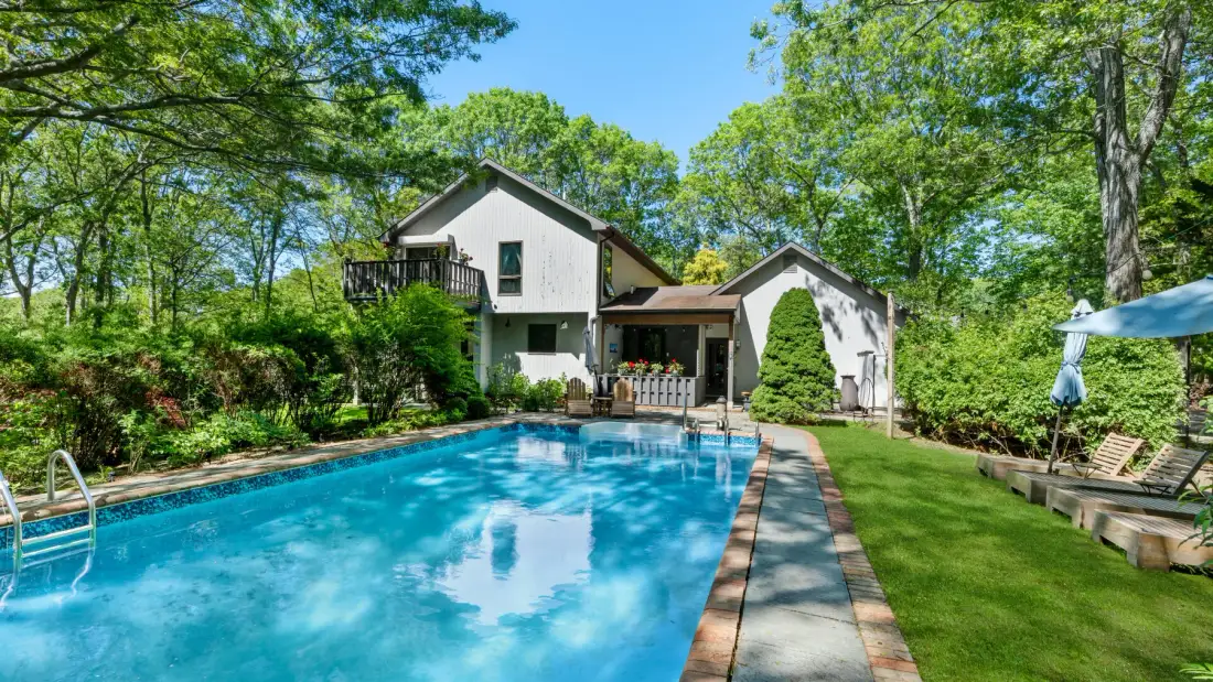 Una piscina exterior está junto a una casa blanca, un césped y árboles frondosos.