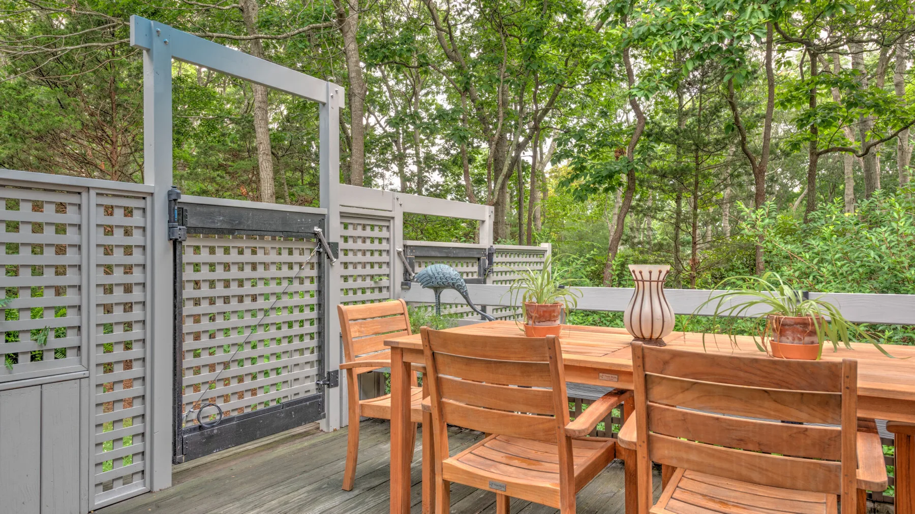 Una mesa de comedor de madera y asientos al aire libre junto a un patio cercado y frondoso.