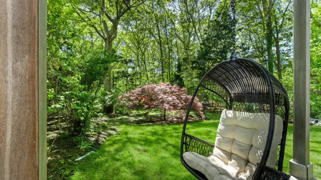 Un jardín con una silla colgante acolchada bajo árboles frondosos.