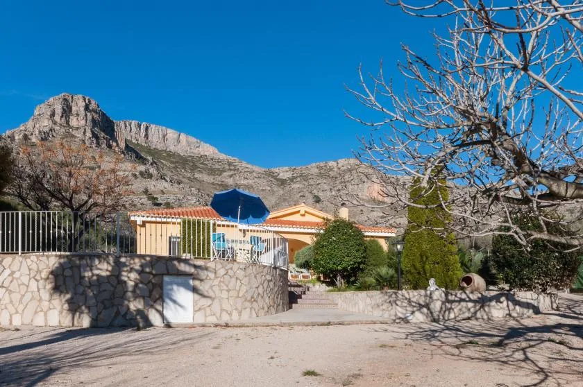 A house and landscaped yard sit beneath a rocky mountain ridge on a sunny day.
