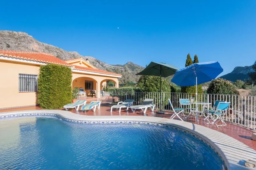 An outdoor swimming pool beside a house, with patio seating and umbrellas.