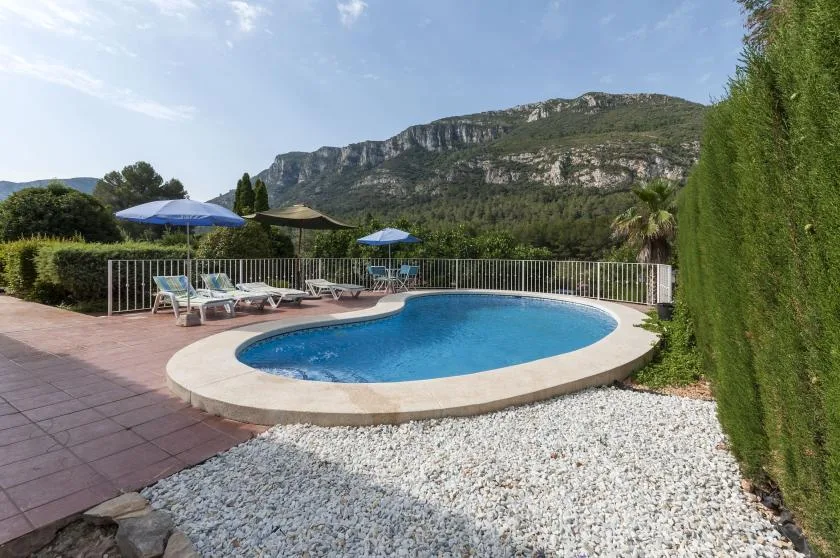 An outdoor swimming pool sits beside a tiled patio, with hills in the background.