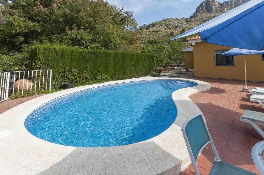 An outdoor swimming pool sits beside a tiled patio with blue shade umbrellas.
