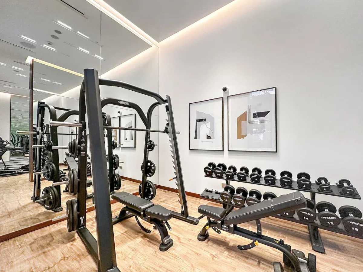 Un gimnasio con equipo de pesas, bancos y mancuernas junto a la pared.