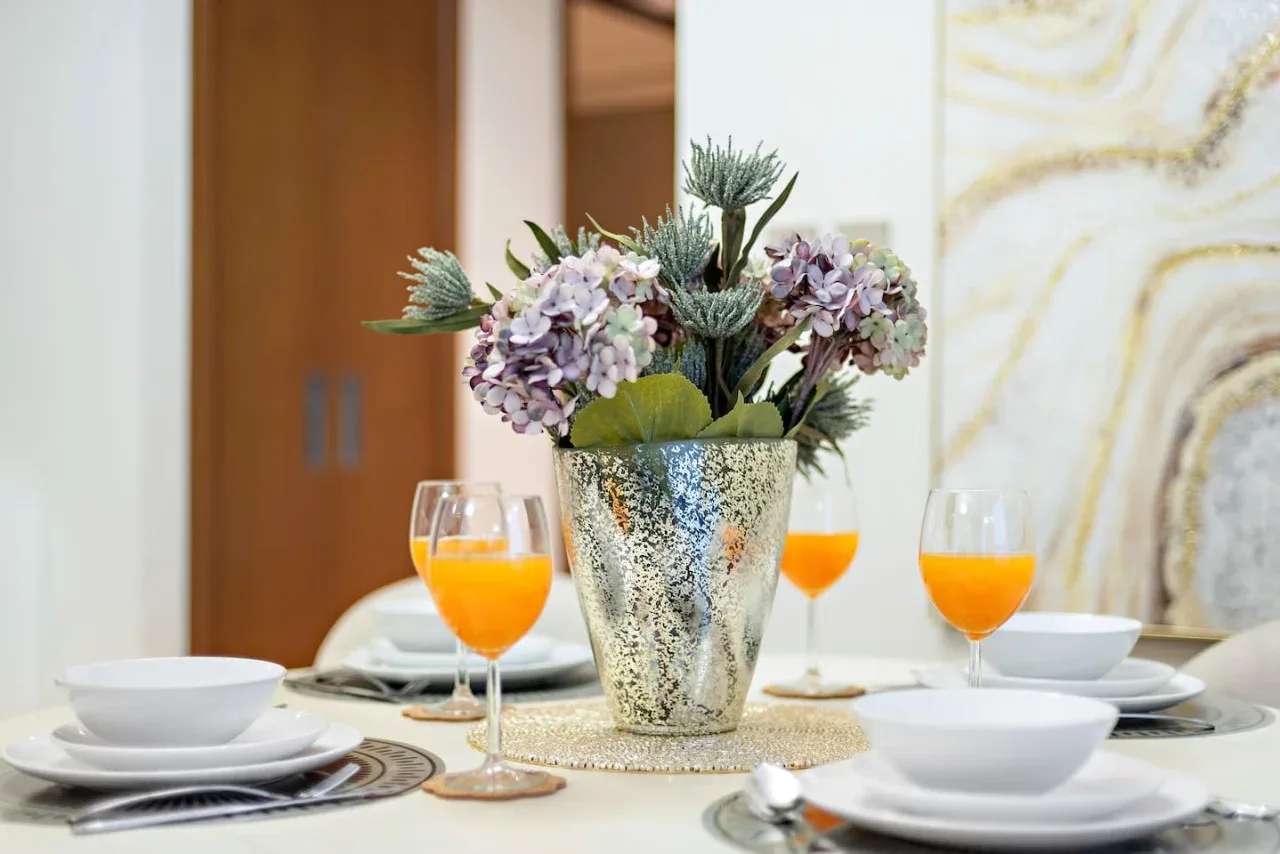 Una mesa de comedor está dispuesta con platos, bebidas naranjas y un arreglo floral.