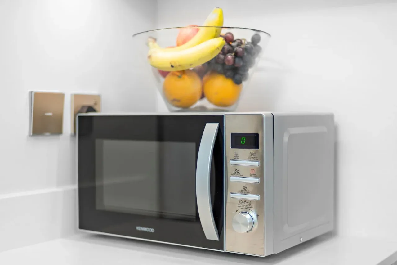 Un microondas está sobre una encimera de cocina, debajo de un recipiente de vidrio con fruta.