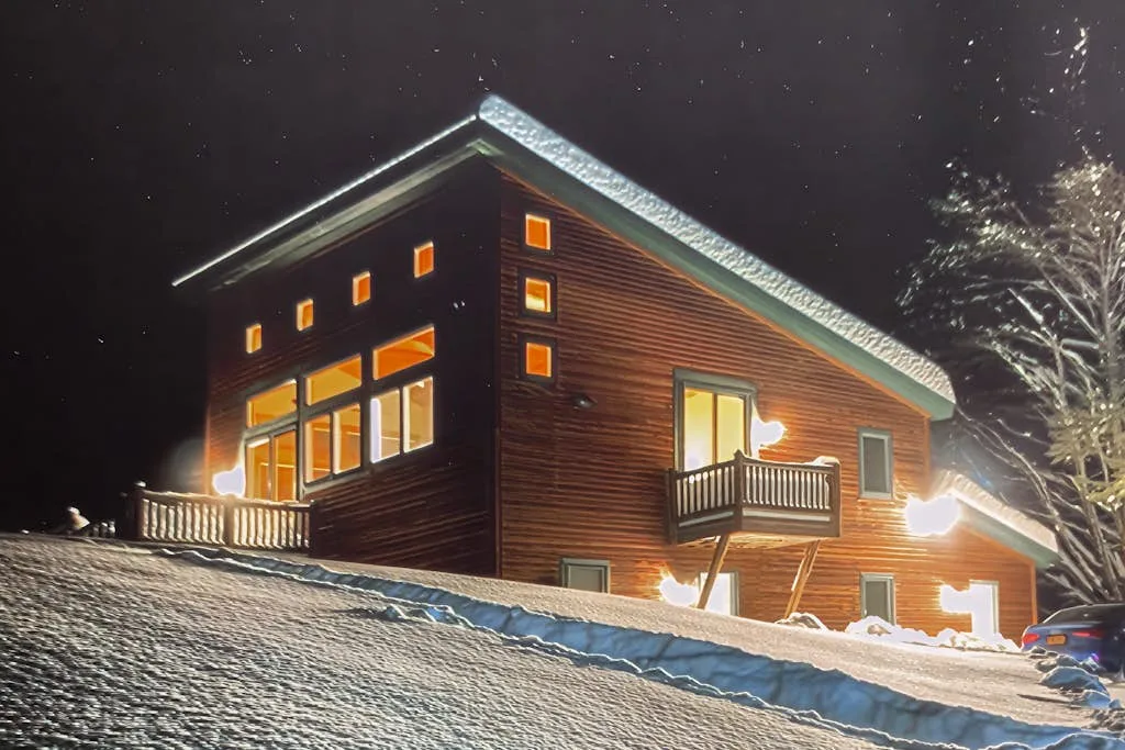 A warmly lit wooden house stands on a snowy slope at night.