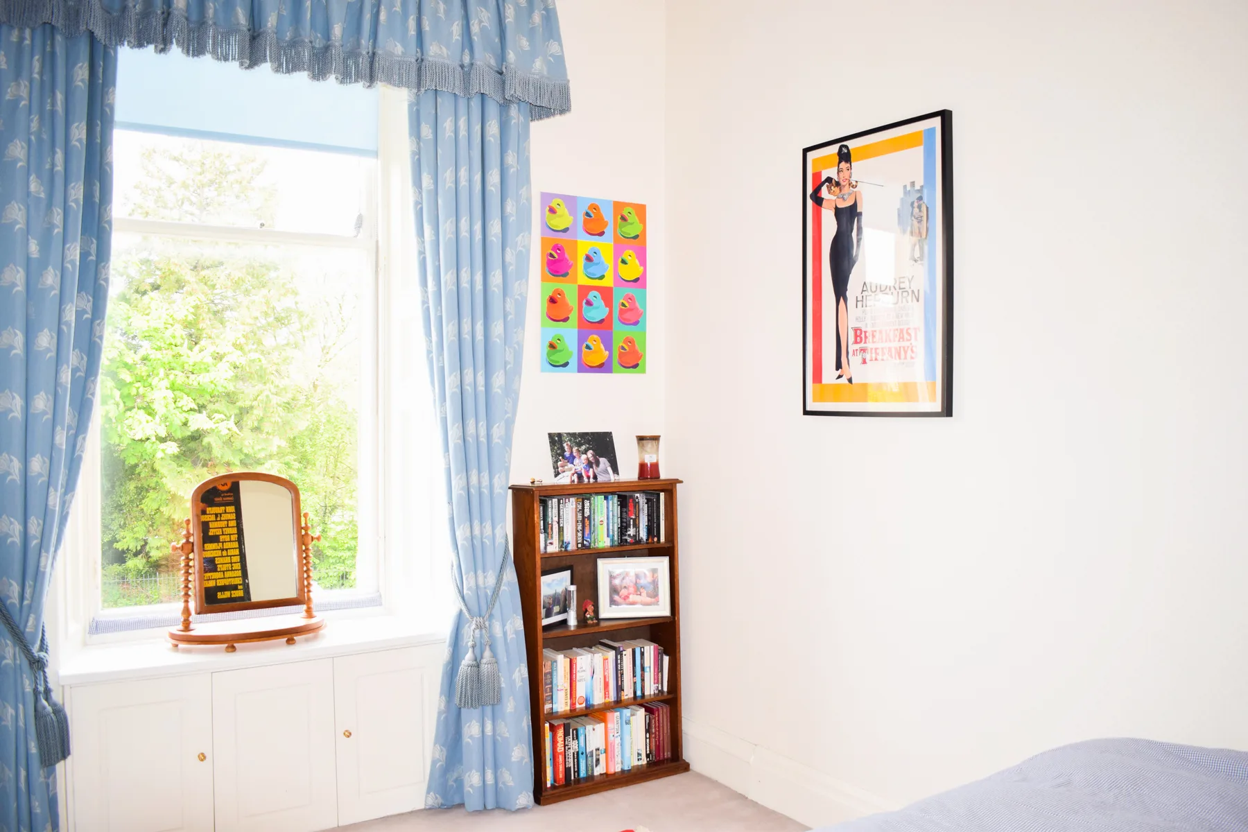 A bright bedroom with blue curtains around a large window and a small bookcase.