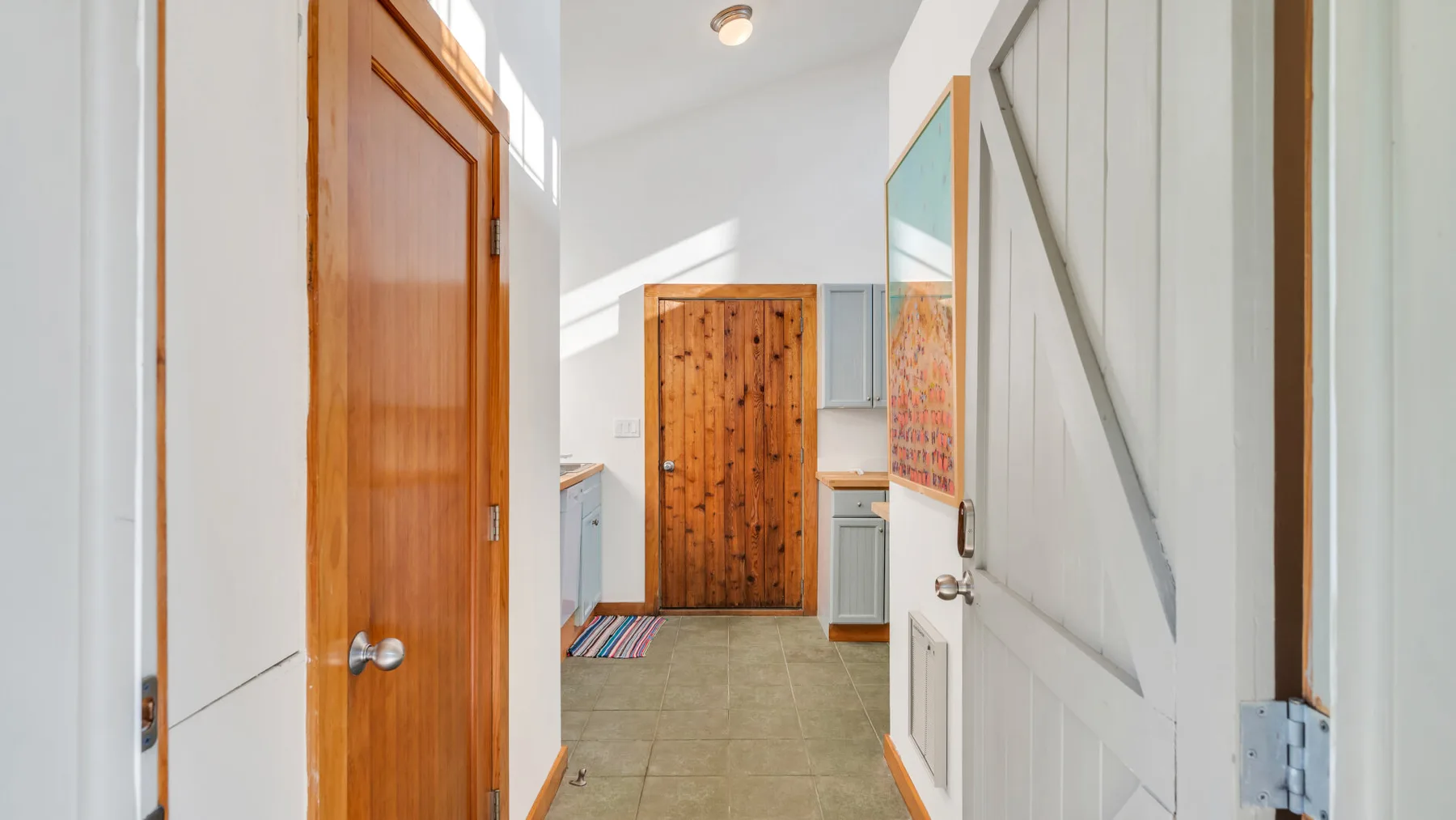 Un pasillo luminoso conduce a una puerta de madera, con una puerta blanca abierta en primer plano.