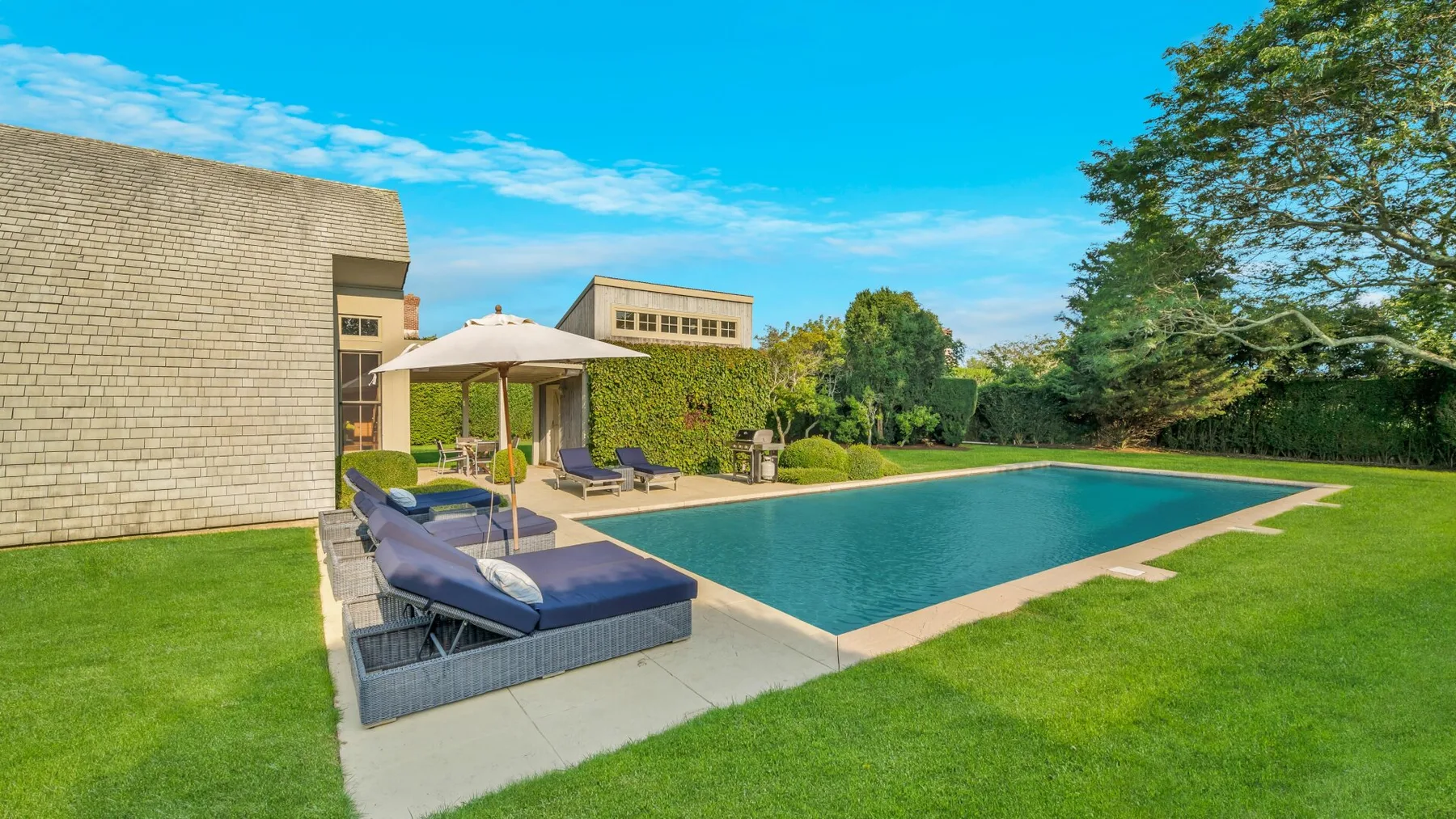 Una piscina al aire libre junto a un patio con asientos reclinables y una sombrilla.