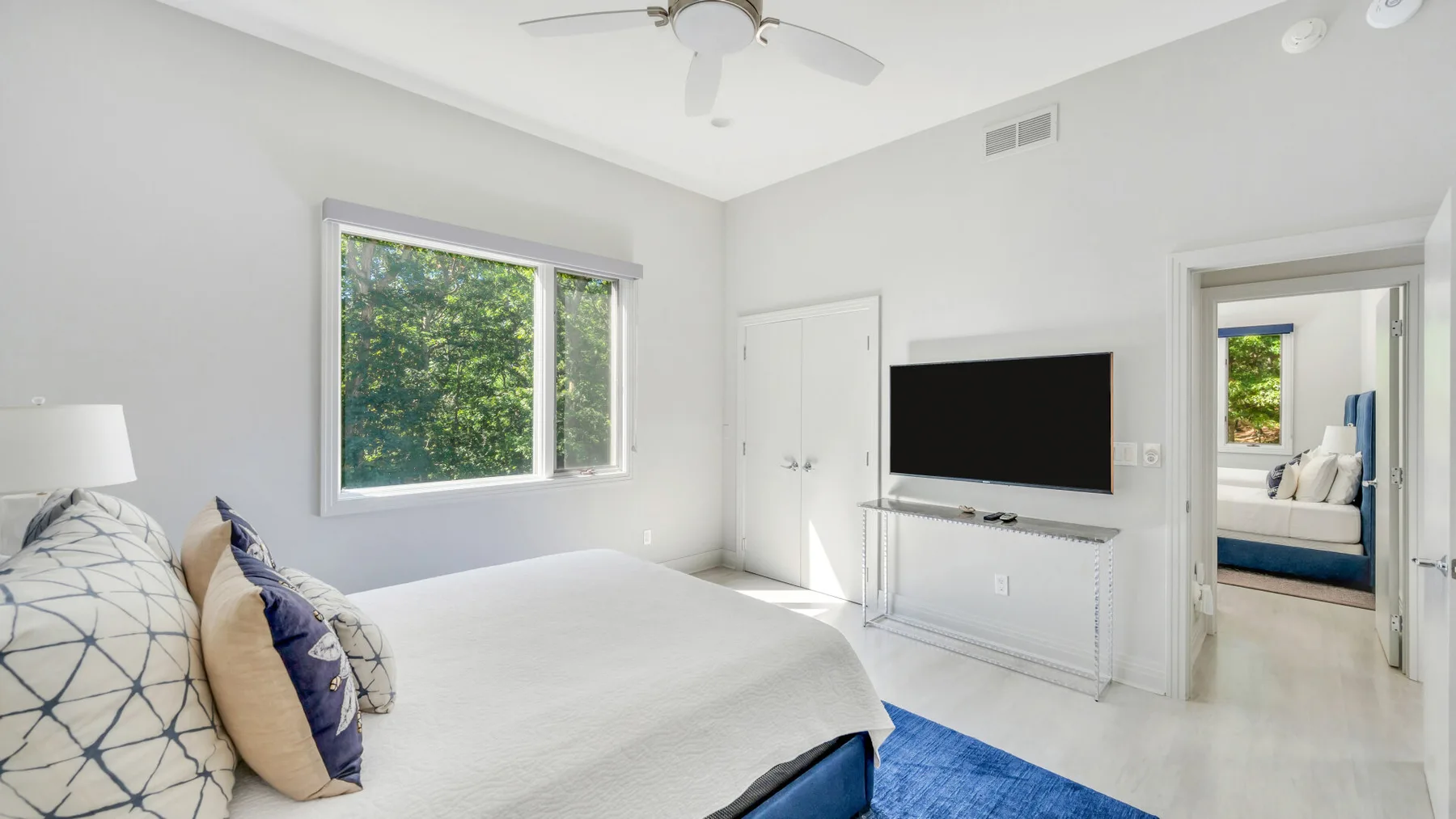 Un dormitorio luminoso con una cama grande, un televisor en la pared y una ventana con vista a la vegetación.