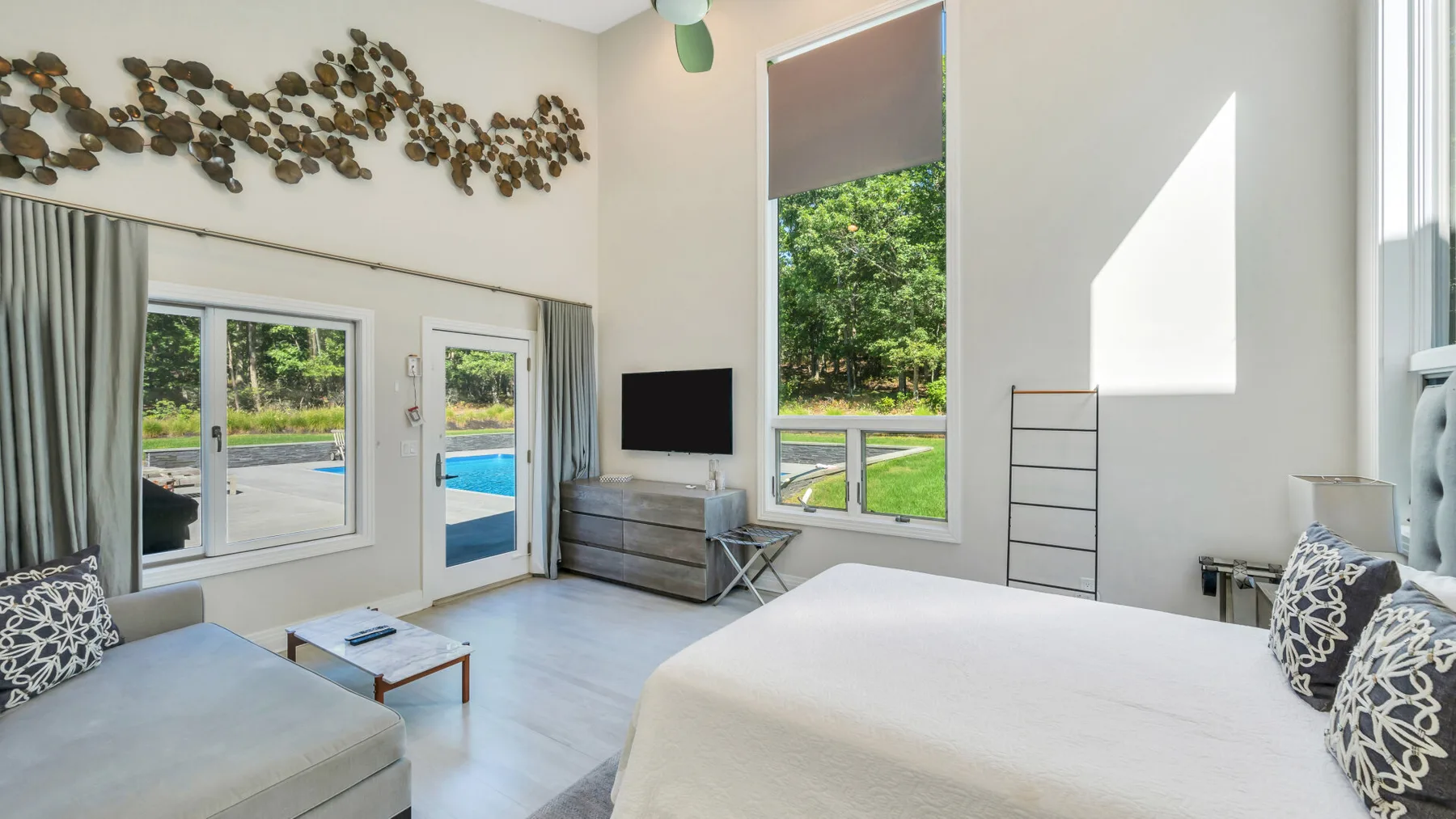 Un dormitorio luminoso con cama, asientos y grandes ventanas con vistas a la vegetación.