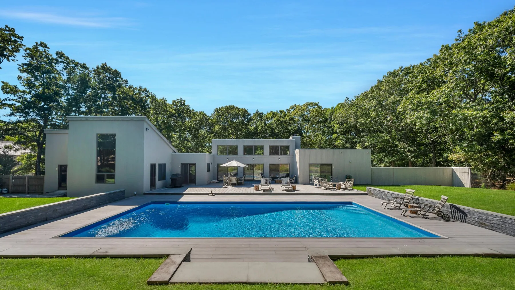 Una piscina y una terraza pavimentada junto a una casa moderna, rodeadas de árboles.