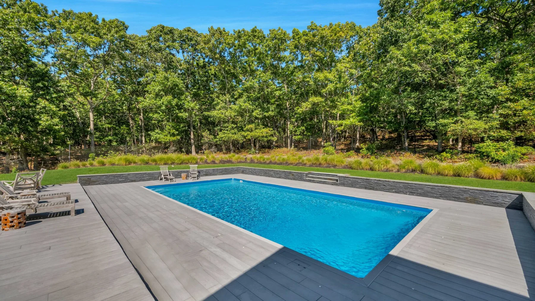 Una piscina al aire libre está rodeada por una amplia terraza gris y árboles.