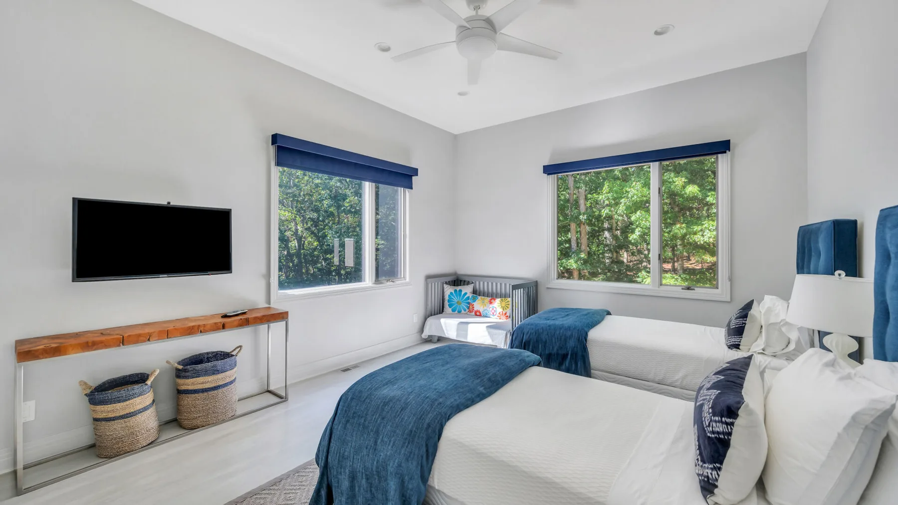 Un dormitorio luminoso con ropa de cama blanca, detalles azules y grandes ventanas con vistas a la vegetación.