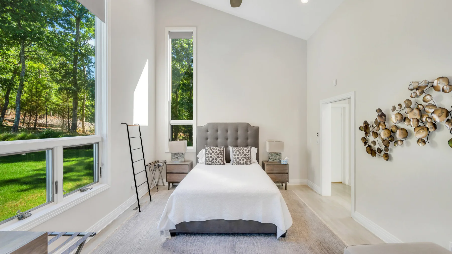 Un dormitorio luminoso con una cama, ventanas altas y vistas a árboles verdes.