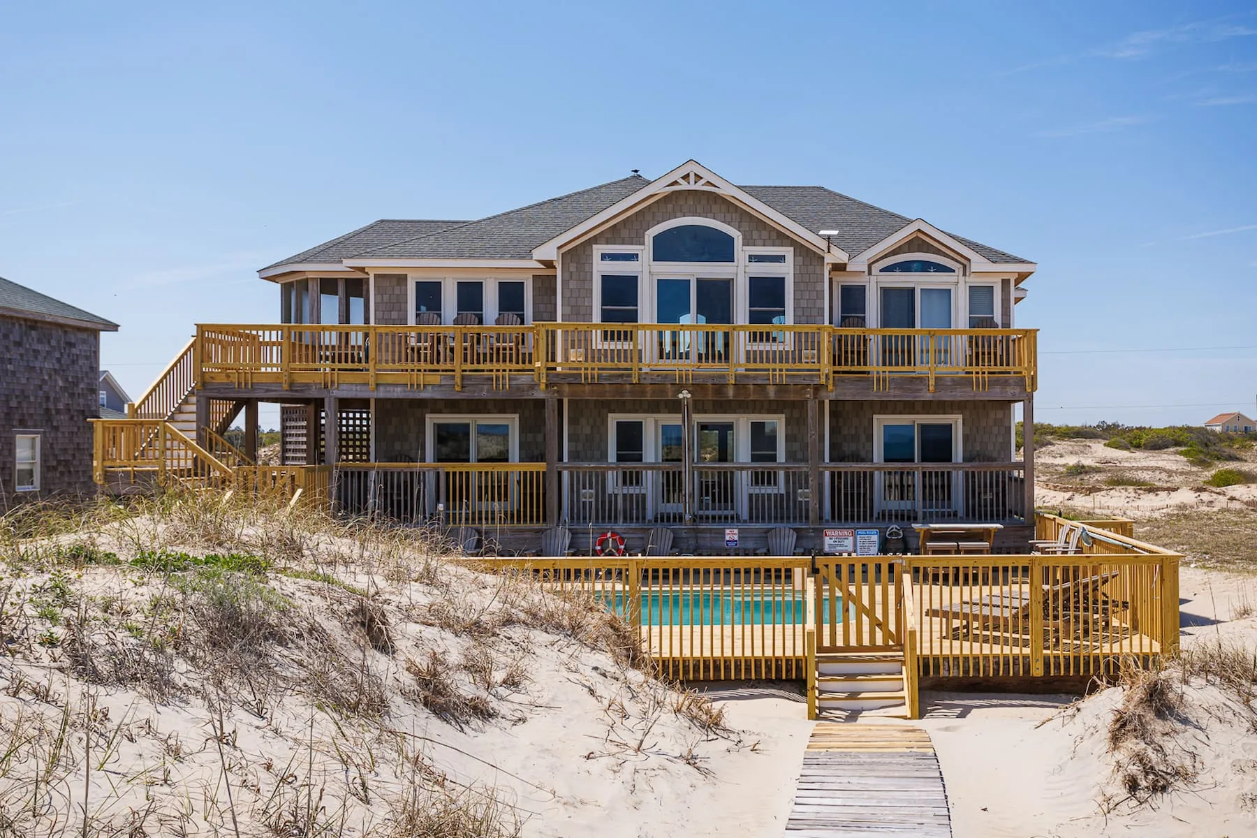 Una gran casa junto a la playa con terrazas de madera y una piscina tras las dunas.