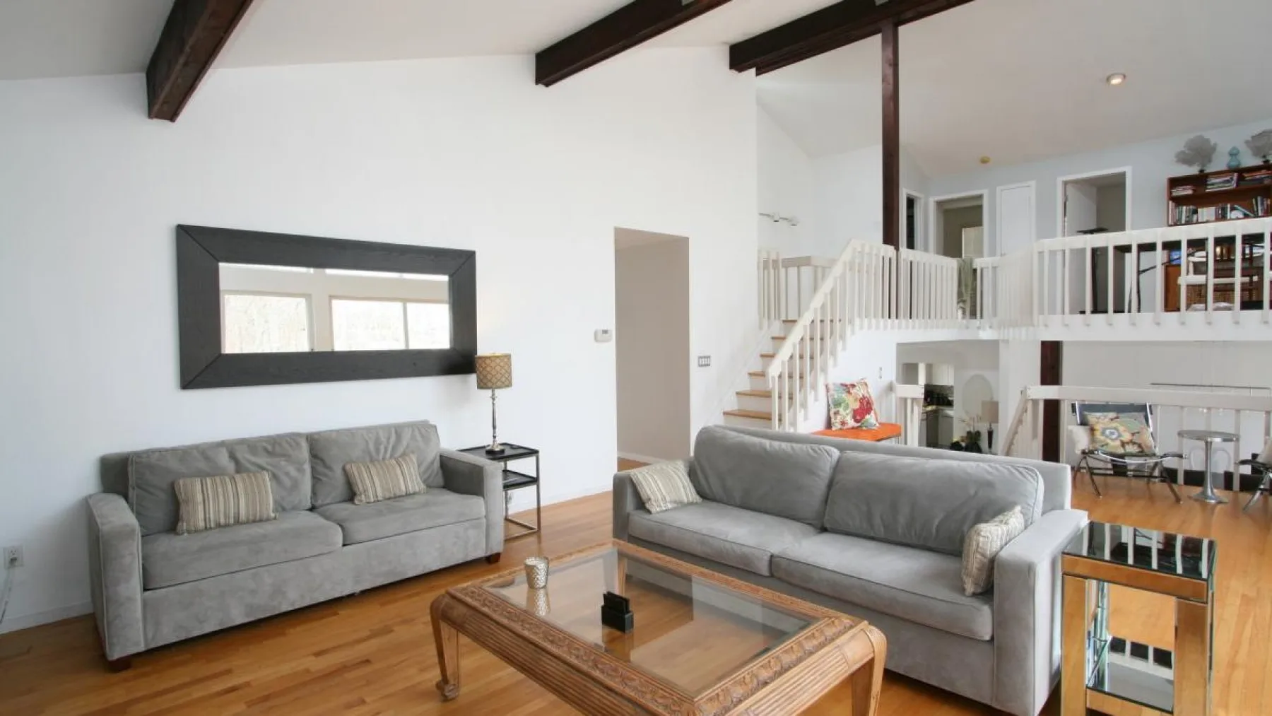 A bright living room with gray seating, a glass coffee table, and a white staircase.