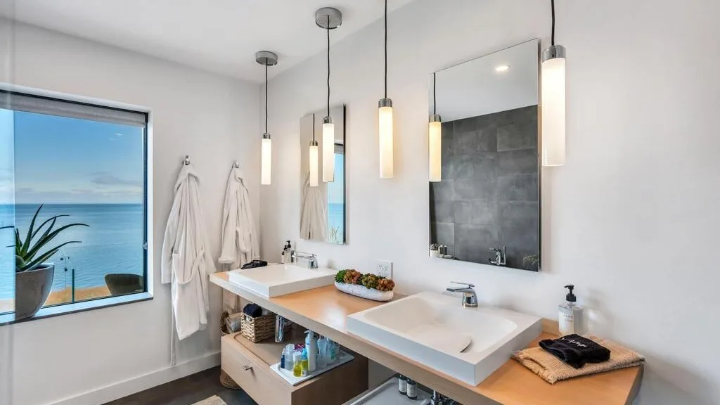 Un tocador de baño luminoso con dos lavabos, lámparas colgantes y una ventana con vistas al agua.