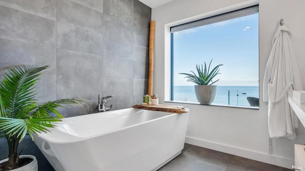 Un baño luminoso tiene una bañera exenta junto a una gran ventana con vistas al mar.