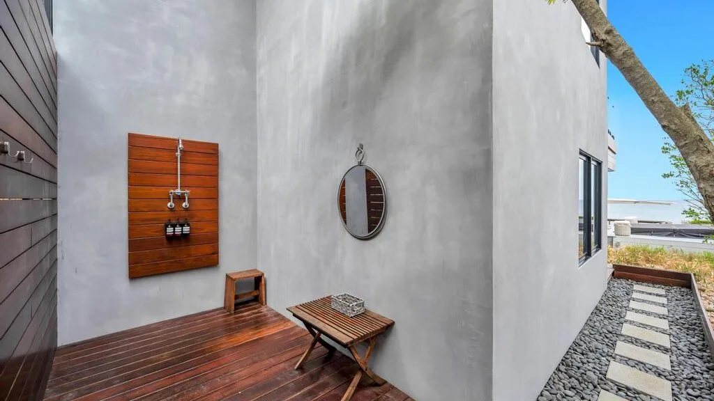 Patio exterior con una terraza de madera, una mesita y un sendero de piedras junto a una pared blanca.