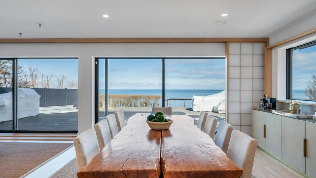 Comedor con una larga mesa de madera junto a grandes ventanas con vista al océano.