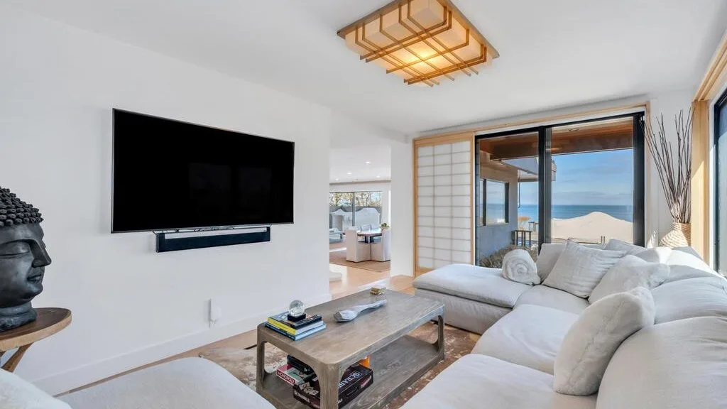 Una sala luminosa con asientos blancos, una mesa de centro y puertas corredizas de vidrio con vista al agua.